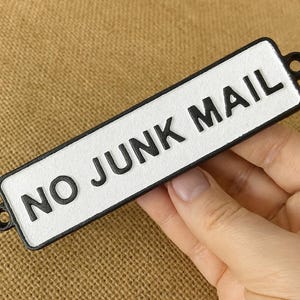 May include: A rectangular sign with the words "NO JUNK MAIL" in black, raised letters. The sign has a white background and a black border. Two small holes are on each end. The sign is held by a hand.