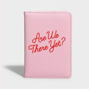 May include: A light pink passport holder with the red text "Are We There Yet?" in a cursive font. The holder is rectangular and appears to be made of leather or a similar material. The edges are stitched.