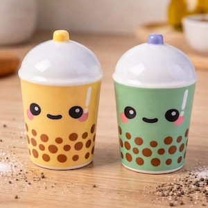 May include: Two ceramic salt and pepper shakers shaped like bubble tea cups. One is yellow with a yellow lid, the other is green with a purple lid. Both have cute faces and brown boba details.