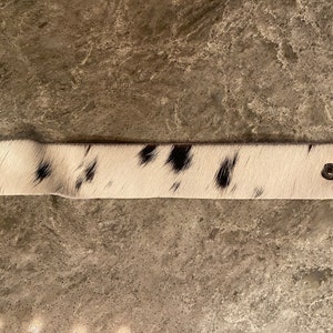 May include: A white and black cowhide leather strap with silver metal accents. The strap is approximately 10 inches long.