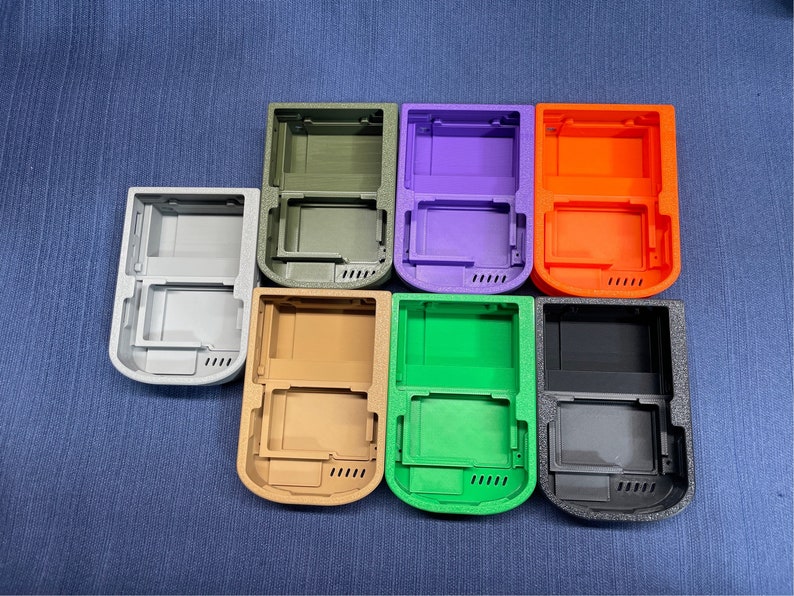 K&ouml;nnte beinhalten: Acht verschiedene farbige Plastikh&uuml;llen f&uuml;r ein Handheld-Spieleger&auml;t. Die H&uuml;llen sind in einem Kreis angeordnet, wobei die Oberseite jeder H&uuml;lle zum Betrachter zeigt. Die Farben der H&uuml;llen sind grau, gr&uuml;n, orange, lila, braun, schwarz und zwei Gr&uuml;nt&ouml;ne.