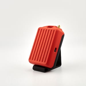 May include: A red and black electronic device with a small antenna on top. The device is sitting on a black stand.