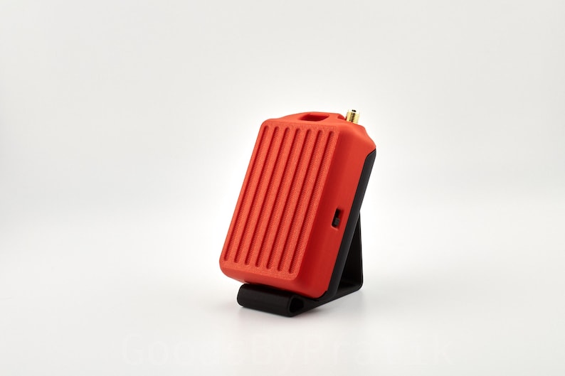 May include: A red and black electronic device with a small antenna on top. The device is sitting on a black stand.
