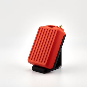 May include: A red and black electronic device with a ribbed surface and a small antenna. The device is sitting on a black stand.