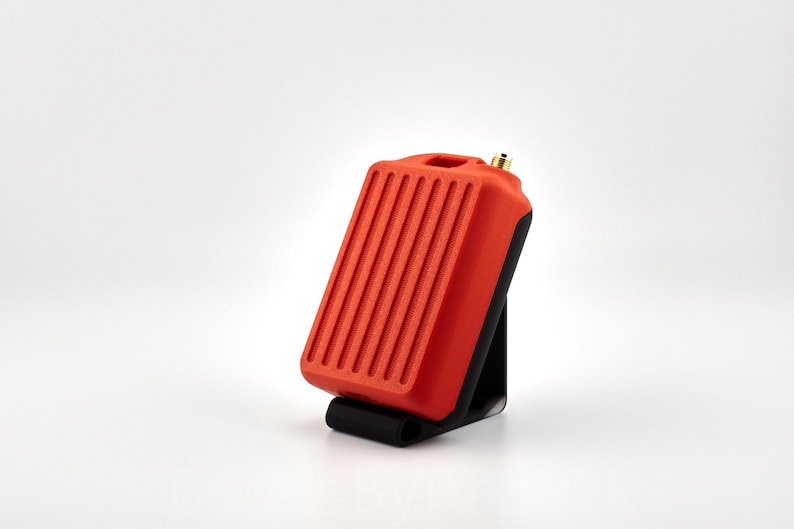 May include: A red and black electronic device with a ribbed surface and a small antenna. The device is sitting on a black stand.
