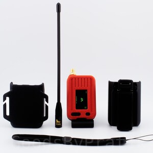 May include: A red handheld radio transmitter with a black antenna, a black wrist strap, and a black carrying case. The radio has a small screen with a green symbol on it.
