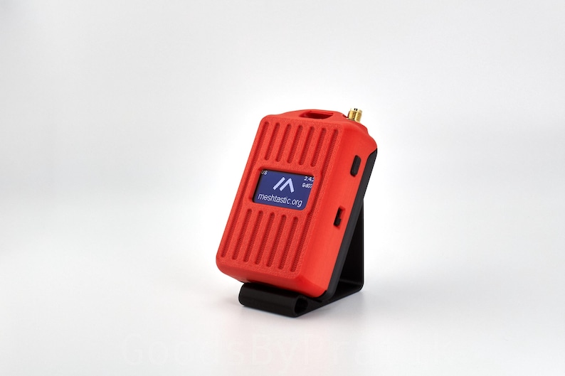 May include: A red mesh network device with a black stand and a small blue screen displaying the text "meshmastic.org" and "2.4.2 dbtc".