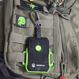 May include: A black and lime green Sensecap electronic device with a central circular button, encased in a protective frame. The device is attached to an olive drab tactical backpack via a black strap and a dark metal carabiner clip.