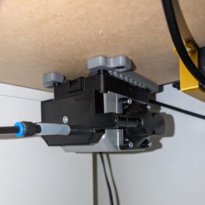 Op de afbeelding: Een zwarte en zilveren luchtpomp is onder een houten oppervlak gemonteerd. De pomp heeft een cilindrisch ontwerp met een doorzichtige buis en een blauwe connector. Grijze montagebeugels bevestigen de pomp aan het hout.
