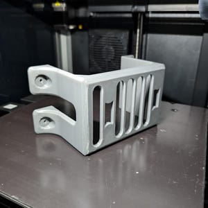 Può includere: Una griglia Jeep stampata in 3D grigio chiaro, con fessure verticali e punti di montaggio. Il pezzo è appoggiato su una superficie scura, probabilmente un piano di stampa 3D, con i componenti interni della stampante visibili.