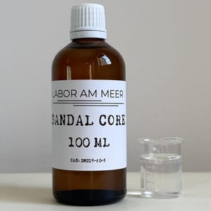 May include: A brown glass bottle with a white cap and a label that reads "LABOR AM MEER SANDAL CORE 100 ML". A small clear beaker is next to the bottle. The liquid in the beaker is clear.