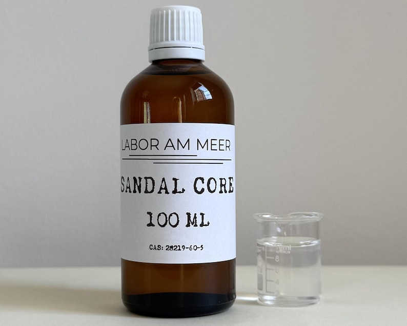 May include: A brown glass bottle with a white cap and a label that reads "LABOR AM MEER SANDAL CORE 100 ML". A small clear beaker is next to the bottle. The liquid in the beaker is clear.