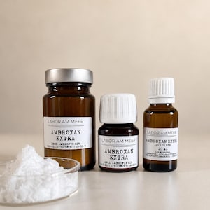 May include: Three amber glass bottles with silver or white caps and white labels reading Labor Am Meer Ambroxan Extra, alongside a clear glass petri dish containing a small pile of white crystalline powder on a neutral surface.
