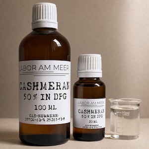 May include: Two amber glass bottles with white caps and white labels reading LABOR AM MEER CASHMERAN 50% IN DPG. The larger bottle is 100 ml and the smaller is 20 ml. A clear plastic measuring cup with 10 ml markings sits next to the smaller bottle.