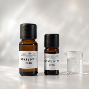 May include: Two amber glass bottles with black caps and white labels reading Labor Am Meer Ambrettolide, one in 20 ml and one in 10 ml, positioned next to a small clear glass measuring beaker with 10 ml markings on the side of the container.
