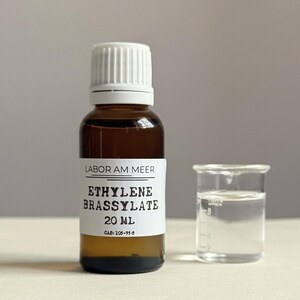 May include: A brown glass bottle with a white cap and a label that reads "ETHYLENE BRASSYLATE 20 ml." Next to it is a small glass beaker with a clear liquid inside. The text "LABOR AM MEER" is also on the label.