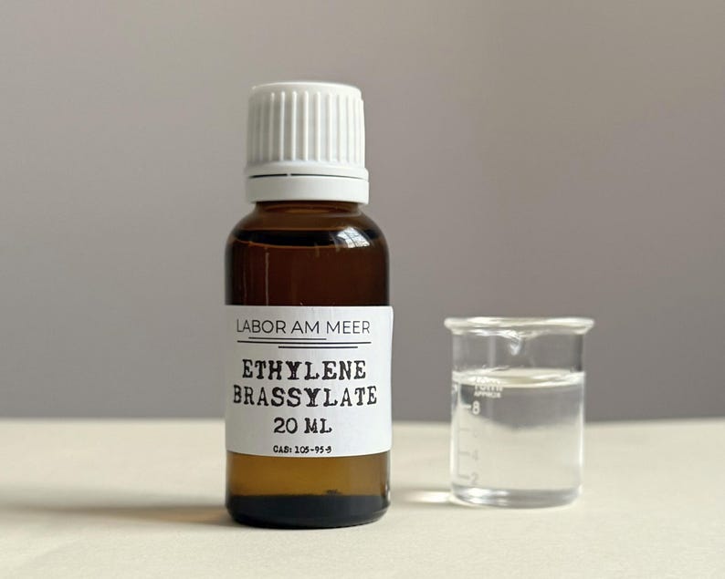 May include: A brown glass bottle with a white cap and a label that reads "ETHYLENE BRASSYLATE 20 ml." Next to it is a small glass beaker with a clear liquid inside. The text "LABOR AM MEER" is also on the label.