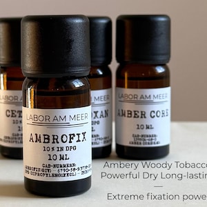 May include: Four small amber glass bottles with black caps are displayed. The bottles have white labels with black text, including "LABOR AM MEER" and "AMBROFIX 10% IN DPG 10 ML." Additional text describes the scent as "Ambery Woody Tobacco" with "Extreme fixation power."