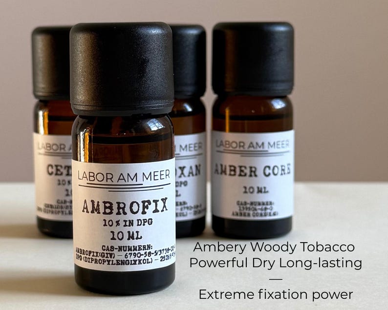 May include: Four small amber glass bottles with black caps are displayed. The bottles have white labels with black text, including "LABOR AM MEER" and "AMBROFIX 10% IN DPG 10 ML." Additional text describes the scent as "Ambery Woody Tobacco" with "Extreme fixation power."
