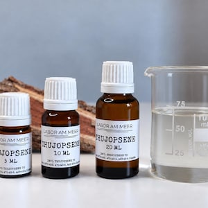 Pode incluir: Três frascos conta-gotas de vidro âmbar rotulados Labor Am Meer Thujopsene nos tamanhos de 5 ml, 10 ml e 20 ml com tampas brancas, posicionados ao lado de um béquer de laboratório de vidro transparente com líquido e marcas de volume visíveis.