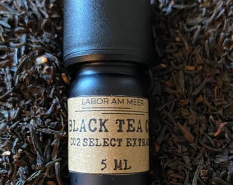 Puede incluir: Una pequeña botella negra con una etiqueta marrón que dice "LABOR AM MEER BLACK TEA CO2 SELECT EXTRACT 5 ML". La botella está sentada sobre un lecho de hojas de té negro sueltas.