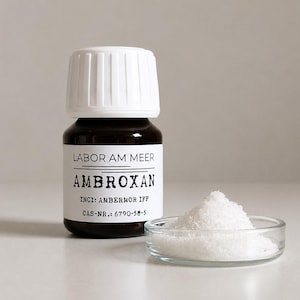 May include: A dark brown glass bottle with a white cap and a label reading LABOR AM MEER AMBROXAN INCI: AMBERMOR IFF CAS-NR.: 6790-58-5, positioned next to a small clear glass dish containing a mound of white crystalline powder substance.