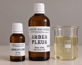 May include: Two amber glass bottles with white caps and labels reading Labor Am Meer Amberfleur, one 20 ml and one 100 ml, alongside a clear glass beaker containing a pale yellow liquid with visible measurement markings up to 100 ml.