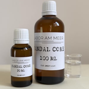 May include: Two amber glass bottles with white labels and caps, labeled "SANDAL CORE" with sizes 20 ml and 100 ml. A small beaker with clear liquid is next to the bottles. The text "LABOR AM MEER" is above the product name.