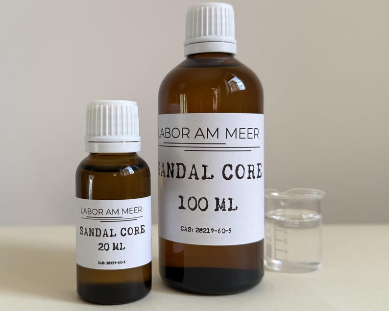 May include: Two amber glass bottles with white labels and caps, labeled "SANDAL CORE" with sizes 20 ml and 100 ml. A small beaker with clear liquid is next to the bottles. The text "LABOR AM MEER" is above the product name.