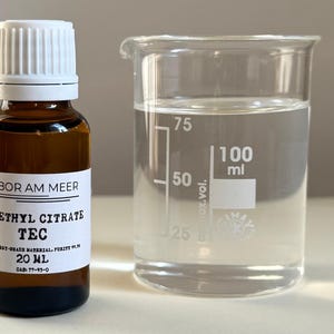 May include: A brown glass bottle with a white cap and label, next to a clear beaker. The label reads "LABOR AM MEER TRIETHYL CITRATE TEC 20 ml". The beaker contains a clear liquid and has volume markings up to 100 ml.