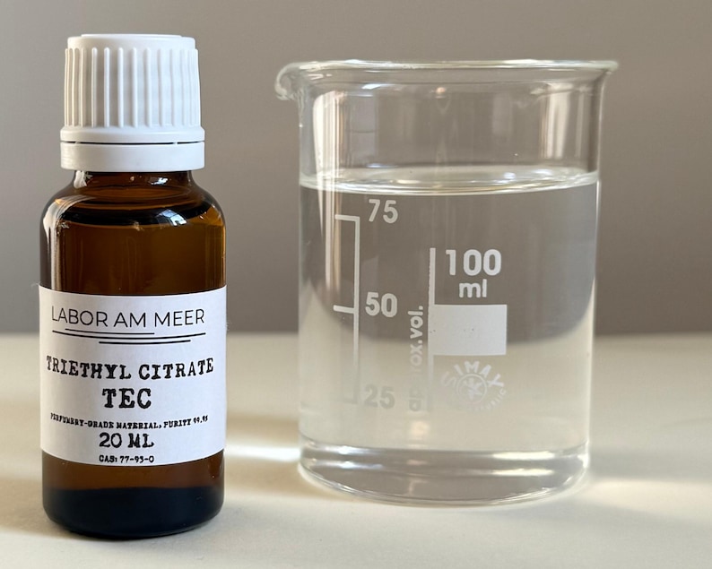 May include: A brown glass bottle with a white cap and label, next to a clear beaker. The label reads "LABOR AM MEER TRIETHYL CITRATE TEC 20 ml". The beaker contains a clear liquid and has volume markings up to 100 ml.