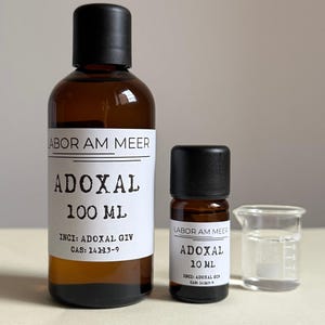 Puede incluir: Dos botellas de vidrio marrón con tapas negras, etiquetadas "ABOR AM MEER" y "ADOXAL". Una botella contiene 100 ml, la otra 10 ml. Un pequeño vaso de precipitados con líquido está junto a la botella más pequeña. Las etiquetas incluyen INCI y CAS.