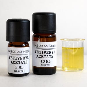 May include: Two amber glass bottles with black lids and white labels. The labels read "LABOR AM MEER VETIVERYL ACETATE" with sizes 5 ml and 10 ml. A small beaker with yellow liquid is next to the bottles.