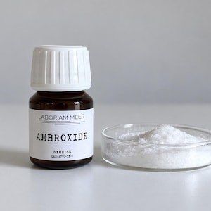 Peut inclure: Un flacon en verre brun avec un bouchon blanc et une étiquette portant l'inscription LABOR AM MEER AMBROXIDE SYMRISE CAS: 6790-58-5, placé à côté d'une boîte de Petri en verre transparent contenant un petit tas de poudre cristalline blanche.