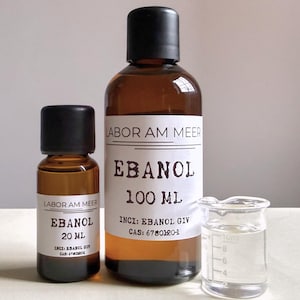 May include: Two amber glass bottles with black caps, labeled "EBANOL" and "LABOR AM MEER". One bottle contains 100 ml, the other 20 ml. A small glass beaker with liquid is next to the bottles. The labels include chemical information.