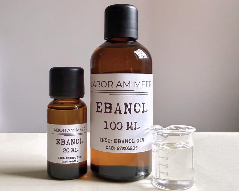 May include: Two amber glass bottles with black caps, labeled "EBANOL" and "LABOR AM MEER". One bottle contains 100 ml, the other 20 ml. A small glass beaker with liquid is next to the bottles. The labels include chemical information.
