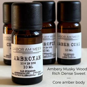 May include: Four amber glass bottles with black caps are displayed. The front bottle has a label that reads "AMBROXAN 10% IN DPG 10 ML" with additional text. The other bottles have labels with the text "LABOR AM MEER" and other text. The image also includes the text "Ambery Musky Woody Rich Dense Sweet Core amber body."
