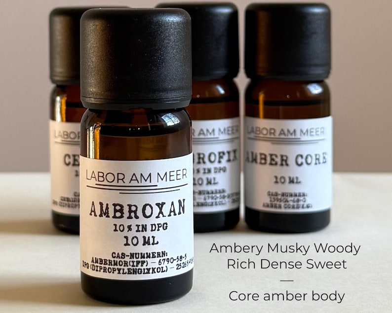 May include: Four amber glass bottles with black caps are displayed. The front bottle has a label that reads "AMBROXAN 10% IN DPG 10 ML" with additional text. The other bottles have labels with the text "LABOR AM MEER" and other text. The image also includes the text "Ambery Musky Woody Rich Dense Sweet Core amber body."