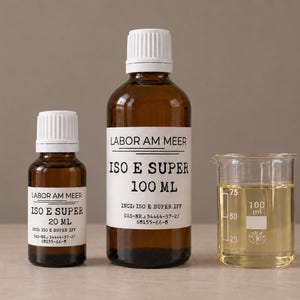 May include: Two amber glass bottles with white caps and white labels reading Labor Am Meer Iso E Super, one 20 ml and one 100 ml, alongside a clear glass beaker containing a pale yellow liquid with measurement markings up to 100 ml.