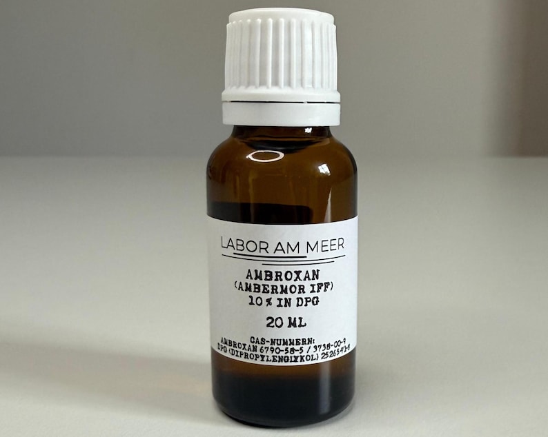 May include: A brown glass bottle with a white cap and a white label. The label reads "LABOR AM MEER" and "AMBROXAN (AMBERMOR IFF) 10% IN DPG 20 mL". The bottle contains a dark liquid.