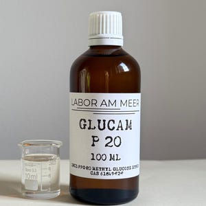 May include: A brown glass bottle with a white cap and a clear glass beaker. The bottle has a white label with black text that reads "GLUCAM P 20 100 ML". The beaker is next to the bottle and contains a clear liquid.