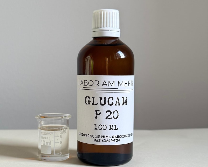May include: A brown glass bottle with a white cap and a clear glass beaker. The bottle has a white label with black text that reads "GLUCAM P 20 100 ML". The beaker is next to the bottle and contains a clear liquid.