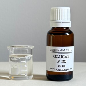 May include: A small glass beaker with liquid next to a brown glass bottle with a white cap. The bottle has a label that reads "LABOR AM MEER GLUCAM P 20 20 ml". The beaker has measurements in milliliters.