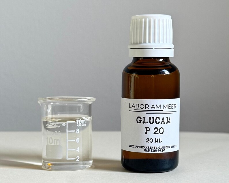 May include: A small glass beaker with liquid next to a brown glass bottle with a white cap. The bottle has a label that reads "LABOR AM MEER GLUCAM P 20 20 ml". The beaker has measurements in milliliters.