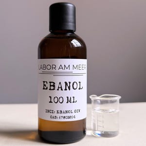 May include: A brown glass bottle with a black cap and a white label that reads "LABOR AM MEER EBANOL 100 ML". A small glass beaker with liquid is next to the bottle.