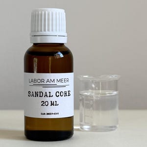 May include: A brown glass bottle with a white cap and label, next to a small clear beaker. The label reads "LABOR AM MEER SANDAL CORE 20 mL". The bottle contains a liquid, and the beaker holds a clear liquid. The background is a neutral color.