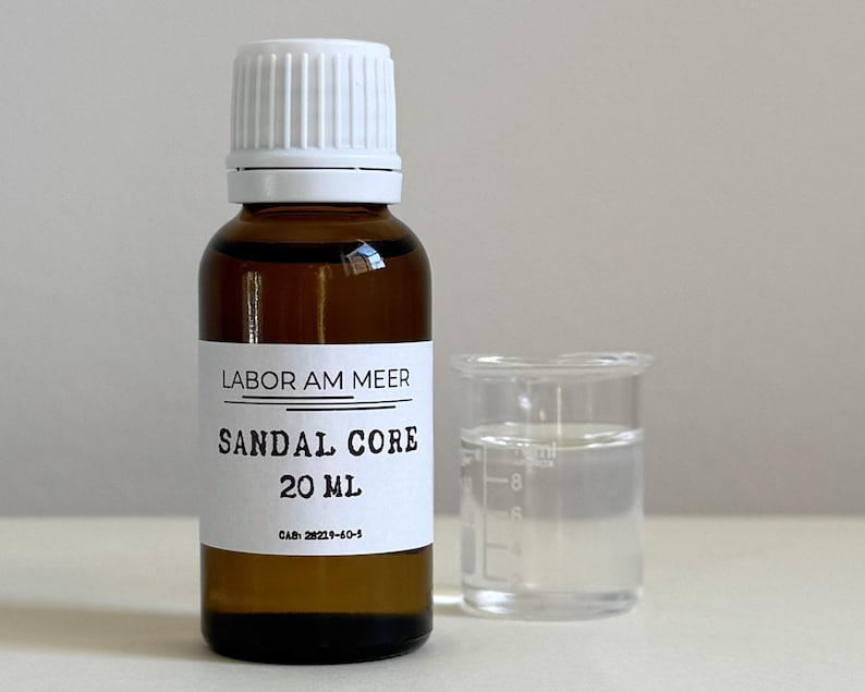 May include: A brown glass bottle with a white cap and label, next to a small clear beaker. The label reads "LABOR AM MEER SANDAL CORE 20 mL". The bottle contains a liquid, and the beaker holds a clear liquid. The background is a neutral color.