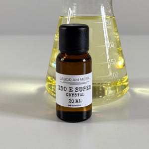Może przedstawiać: Mała, brązowa szklana butelka z czarną zakrętką i białą etykietą z napisem "ISO E SUPER CRYSTAL 20 ml". Znajduje się przed przezroczystym szklanym kolbą wypełnioną ż&oacute;łtym płynem. Na etykiecie widnieje r&oacute;wnież napis "LABOR AM MEER".