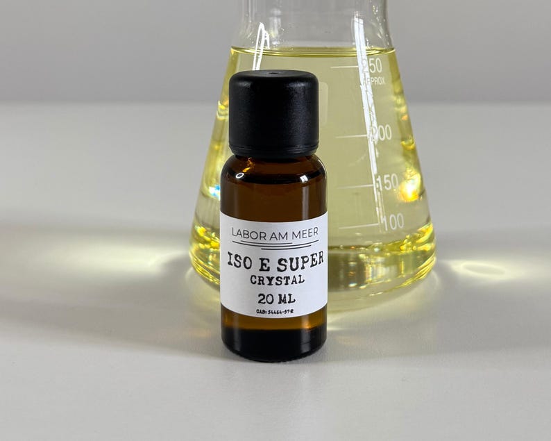 Może przedstawiać: Mała, brązowa szklana butelka z czarną zakrętką i białą etykietą z napisem "ISO E SUPER CRYSTAL 20 ml". Znajduje się przed przezroczystym szklanym kolbą wypełnioną ż&oacute;łtym płynem. Na etykiecie widnieje r&oacute;wnież napis "LABOR AM MEER".