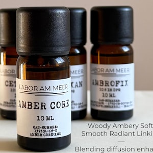 May include: Four small amber glass bottles with black caps. The front bottle has a label that reads "AMBER CORE 10 ML". The text "LABOR AM MEER" is also visible. The image includes the text "Woody Ambery Soft Smooth Radiant Linking".